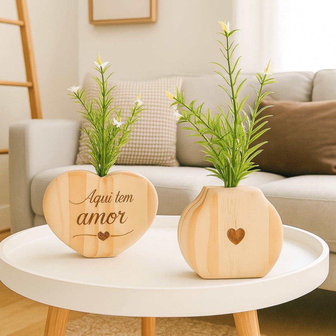 Vaso em formato de coração com frase "Aqui tem amor" ao lado de mini vaso com coração vazado, sobre mesa branca em sala iluminada

