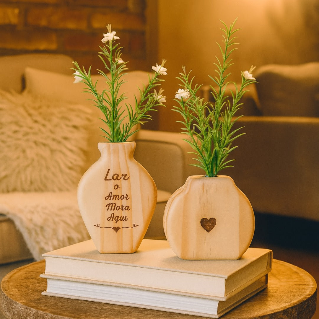 Vaso em formato de garrafinha com frase "Lar o Amor Mora Aqui" ao lado de vaso com coração entalhado, sobre livros decorativos em ambiente aconchegante

