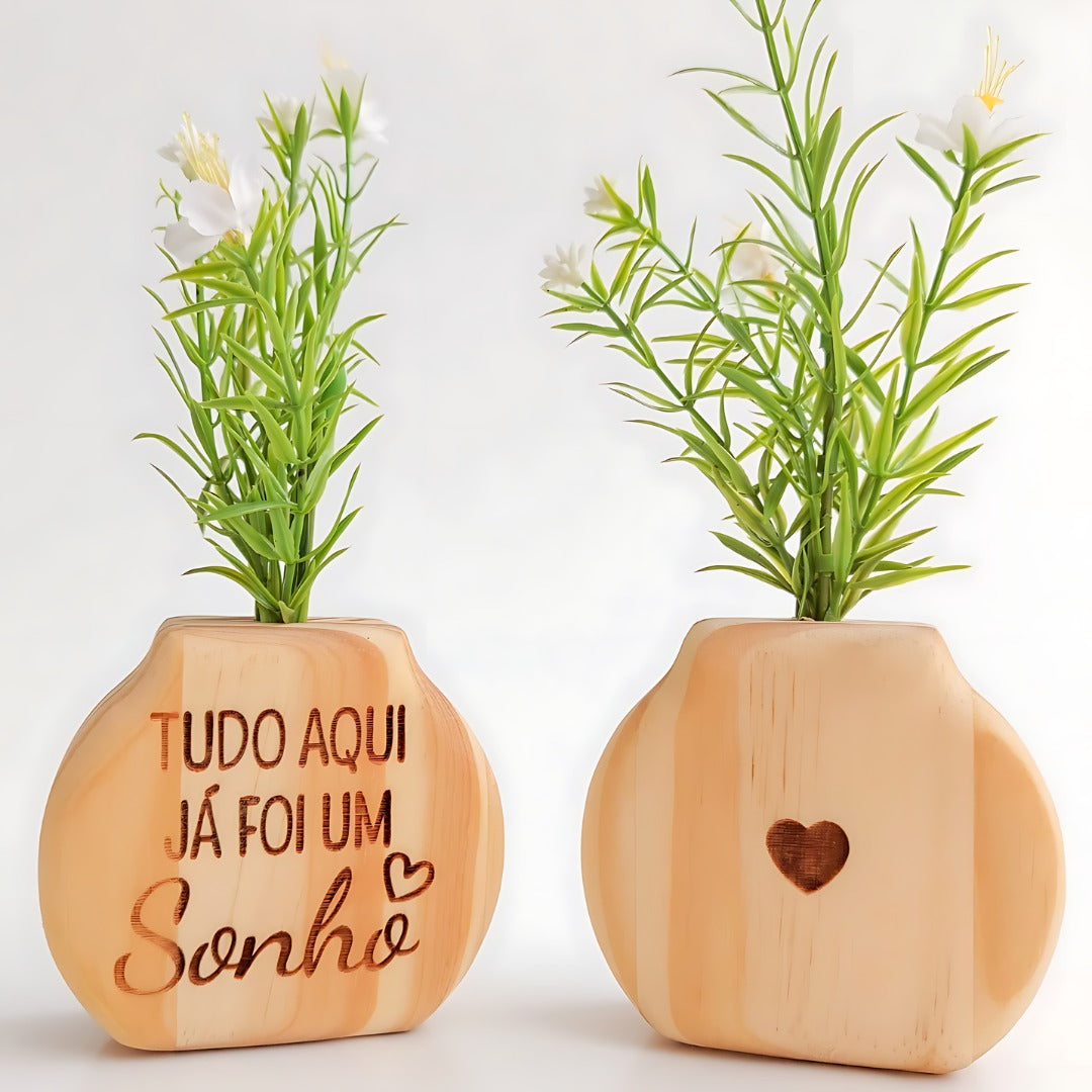 Vasos de madeira clara com flores artificiais, um com frase inspiradora e outro com coração entalhado, em fundo branco

